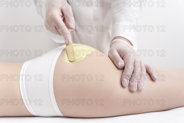 Unrecognizable beautician in gloves applying green hot wax on buttocks of slim woman using spatula.