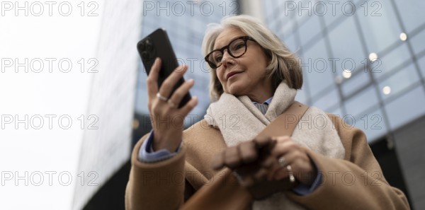 A mature woman wearing glasses and a warm coat is focused on her smartphone, standing on the
