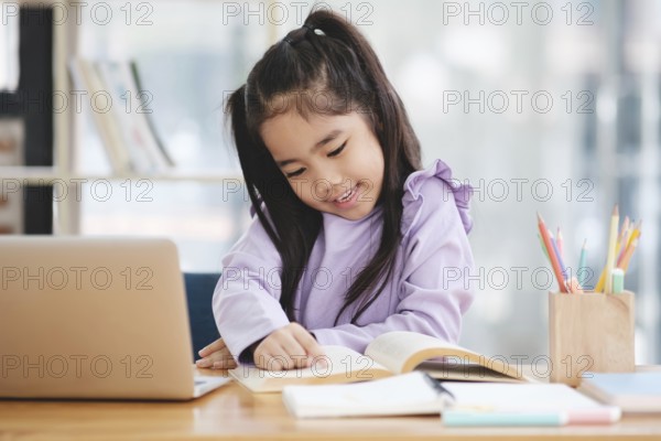 A young girl is sitting at a desk with a laptop and a book. She is smiling and she is enjoying her