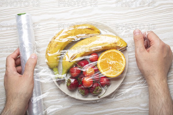 Male hands wrapping assorted different fruit and berries on white plate in plastic clear film top