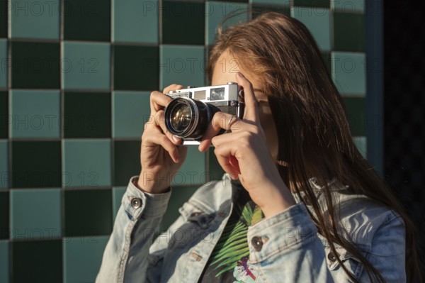 Cool girl model with retro film camera wearing a denim jacket, dark hair outdoors over city wall in
