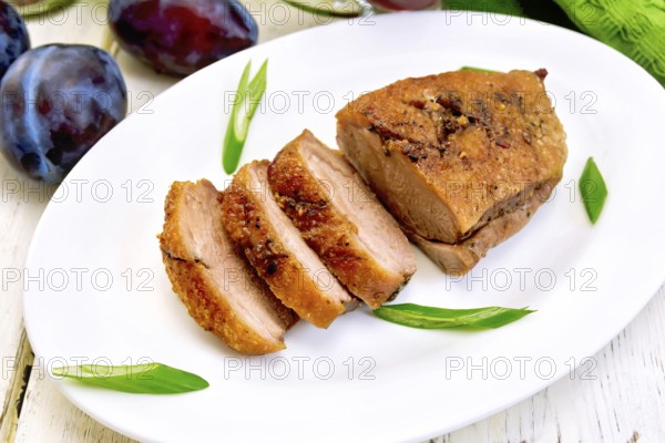 Roasted duck breast with green onions in a white oval plate, towel and plums on a background of