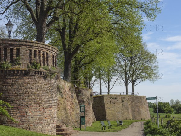 Defence defence tower and brick wall along a walkway, flanked by trees and park benches under a