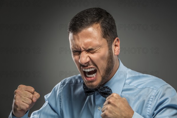 Portrait of angry upset young man in blue shirt and butterfly tie with fists up yelling isolated on