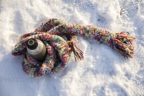 A long, colorful wool scarf lies on the snow. The scarf is wrapped around a thermos. The thermos is