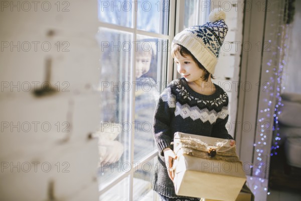 Child sitting at the window on a sunny Christmas day, making presents wrapped in boxes in paper.