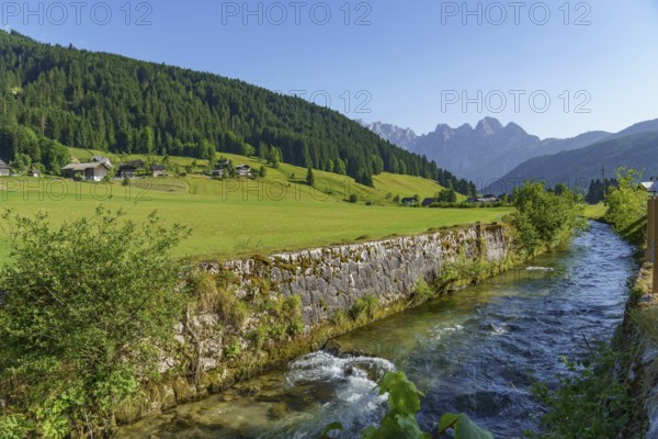 A clear river flows between wide green meadows and a mountain landscape under a bright blue sky,
