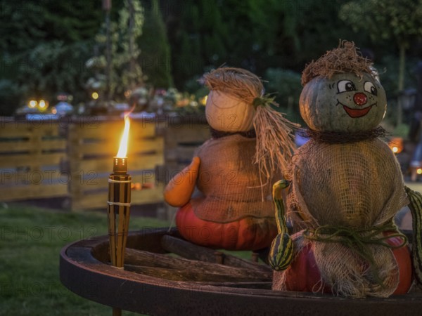 Two pumpkin sculptures sitting on a wooden wheel, illuminated by a burning torch in the evening