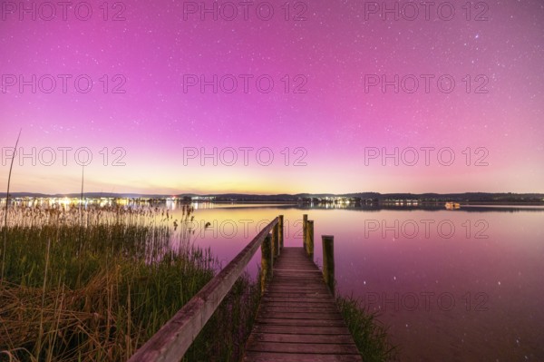 A wooden footbridge leads over a calm lake under a sky with aurora borealis, Reichenau, Lake