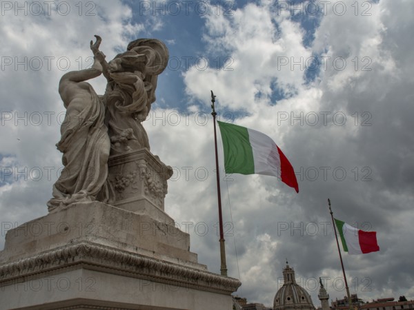 A statue and an Italian flag stand against a dramatic backdrop of clouds with ancient architecture