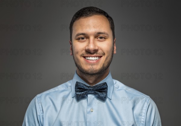 Portrait of happy smiling young man in blue shirt and butterfly tie isolated over gray studio