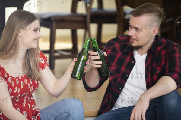 Smiling young people drink beer at a house party. They tap green bottles and look at each other. A