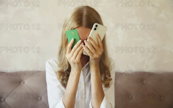Desperate upset young woman holding mobile phone and bank card touching her head and covering face,