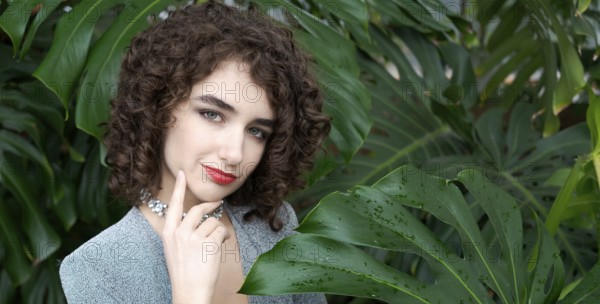 Woman with curly hair is standing in front of a green plant. She is wearing a blue dress and a