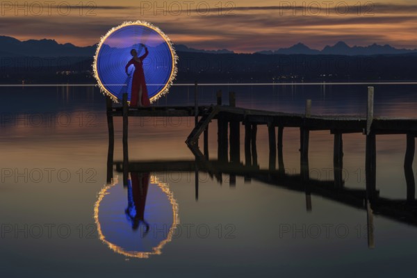 Woman in a red dress on a jetty at sunset on a lake, standing in front of a circle of light created