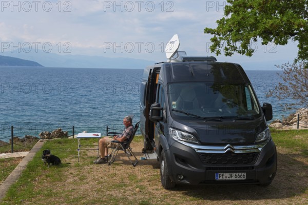 A campsite by the sea, a man sits next to a motorhome and enjoys the view with a dog, Glavotok, Krk