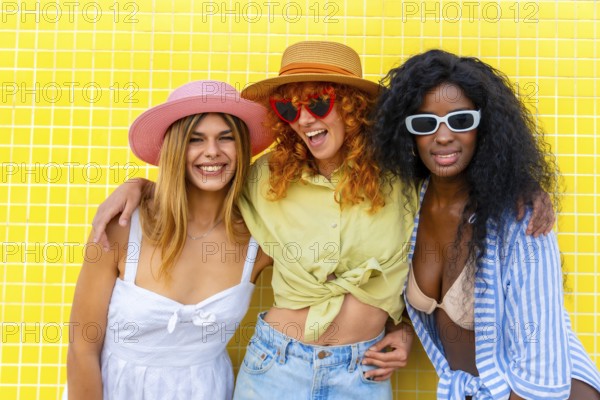 Three cheerful diverse women with colorful hats and sunglasses are posing together against a bright
