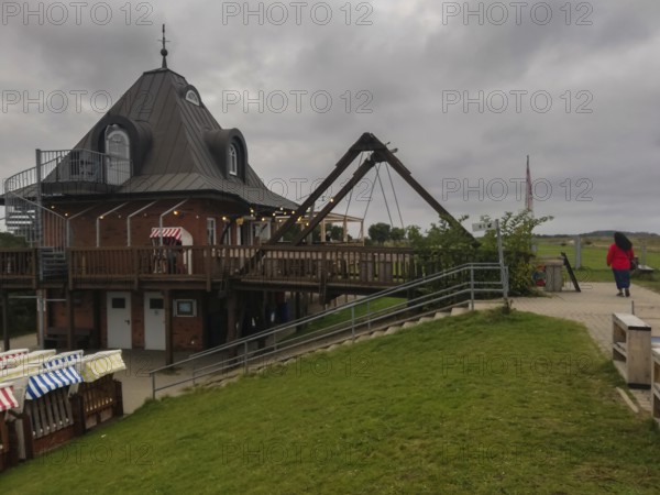 Wooden building with outdoor terrace under a cloudy sky, a person walks past Strandbude Ostseeinsel