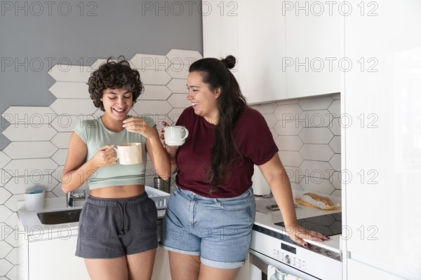 A lesbian couple stands in a cozy kitchen, sipping morning coffee. The modern decor and soft