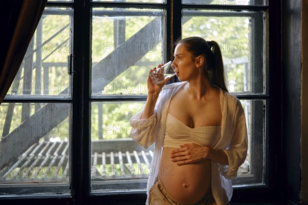 A pregnant woman stands by a large window in her home, sipping water from a glass. She appears