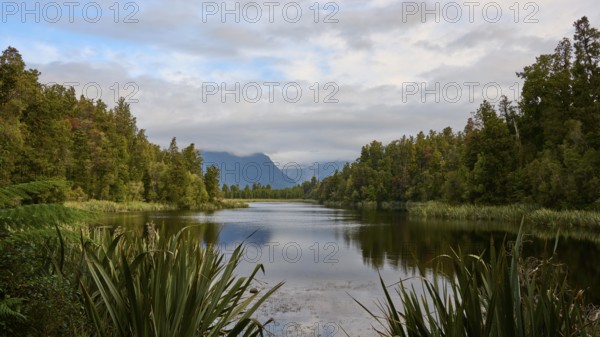 A tranquil lake surrounded by lush vegetation and mountains, with a slightly cloudy sky, summer,