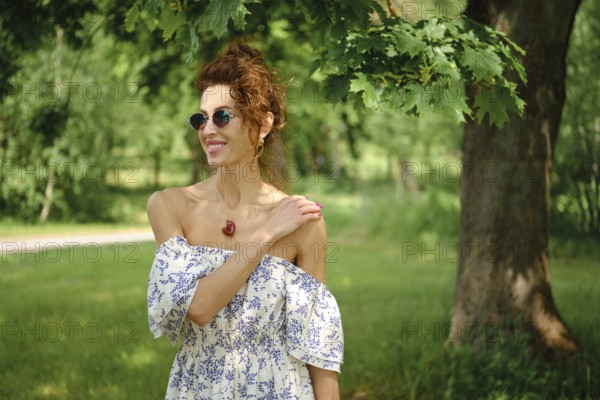 A woman with curly hair stands in a vibrant park, wearing a stylish off-shoulder dress and