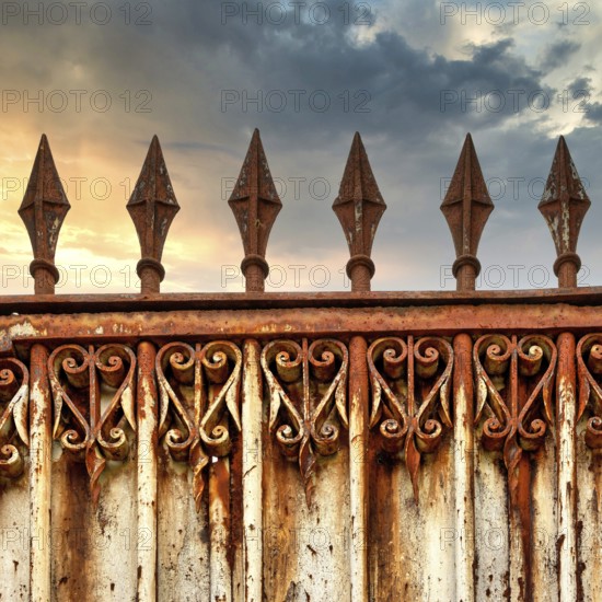 A rusty ornate gate stands tall, featuring intricate designs and pointed spikes. The backdrop shows
