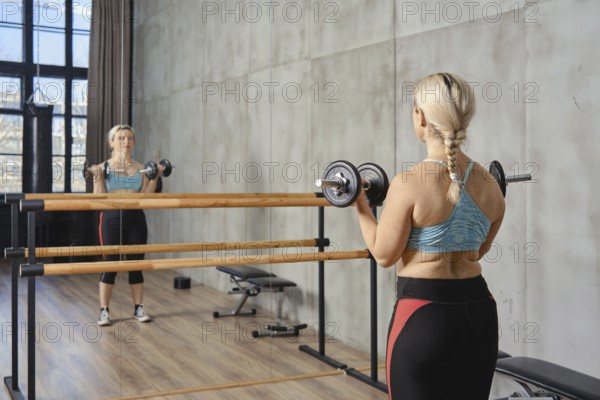 A woman with a braid exercises with dumbbells in a contemporary gym. She is focused, using a mirror