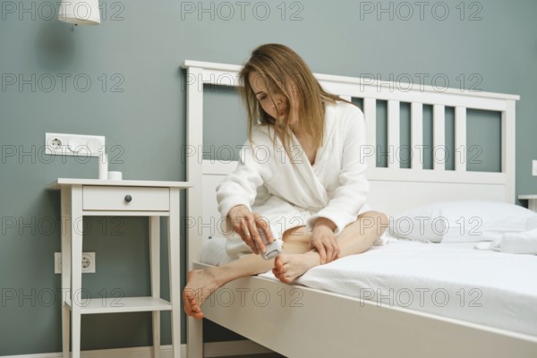 A woman in a bathrobe sits on the edge of a bed using exfoliator on her feet. The room features a