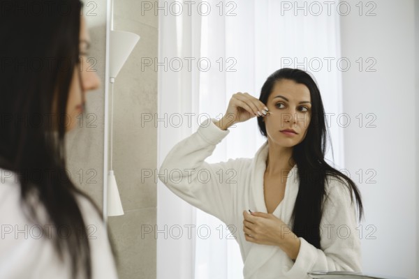 A woman in a white robe looks into a mirror while applying a beauty treatment to her under eye skin