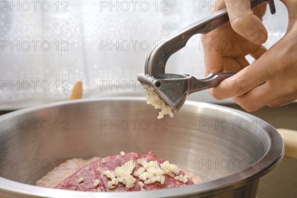 A hand is using a garlic press to add freshly minced garlic to a bowl with ground meat. This