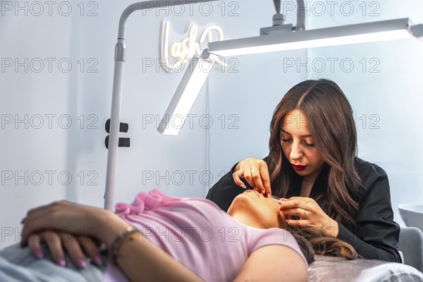 Beautician applying false eyelashes to a client lying on a bed in a beauty salon, illuminated by a