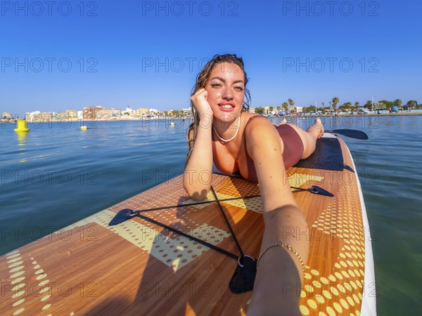 Young woman lying on a paddle surf board, taking a selfie and enjoying a sunny summer day at the