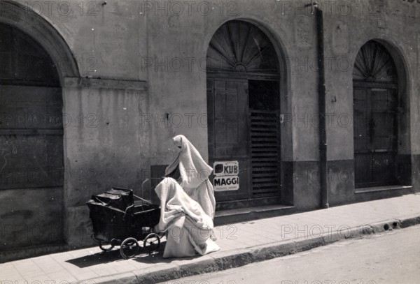 Two Arab women with prams, 1910s, A veiled person sits next to a pram in front of an old building