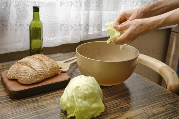 Hands are tearing fresh lettuce over a bowl while a loaf of bread and a bottle of olive oil sit