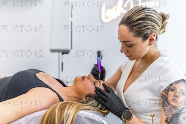 Beautician performing microblading treatment, applying permanent makeup on eyebrows of client lying