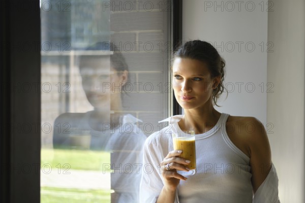 In a stylish, well-lit room, a woman holds a glass of fresh juice. She gazes thoughtfully out the