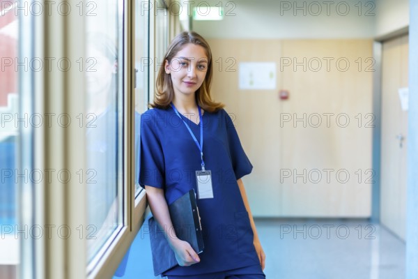 Confident young doctor wearing blue scrubs holding a clipboard while standing in a modern hospital
