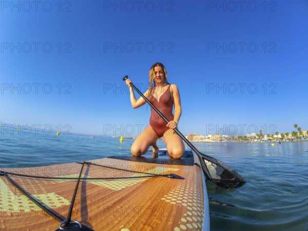 Young woman kneeling on a paddle board, holding a paddle, enjoying a summer watersport activity on