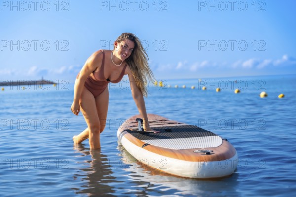 Woman engaging in paddle surfing on a calm summer day, standing beside her paddleboard in clear