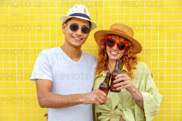 Happy couple toasting with beer bottles while enjoying a summer day against a vibrant yellow tiled