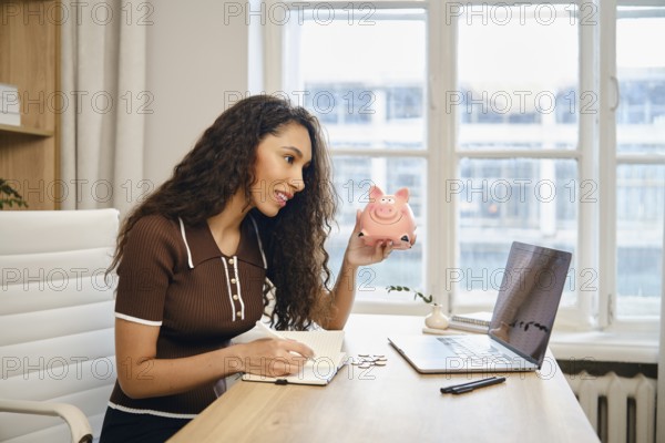 A young woman is actively budgeting at her desk, holding a pink piggy bank in one hand while
