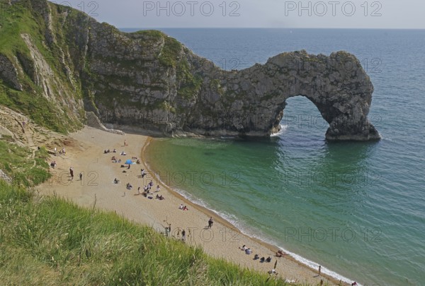 People relaxing on the beach in front of a striking arched cliff and blue sea, Durdle Door,