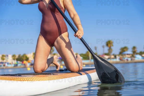 Woman kneeling on a paddleboard, holding a paddle on a calm summer day. Clear blue sky and distant