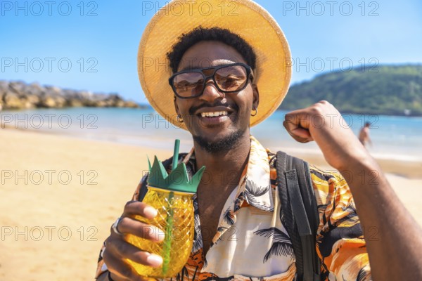 Young man smiling and raising fist while holding a pineapple shaped cocktail on a sunny beach,