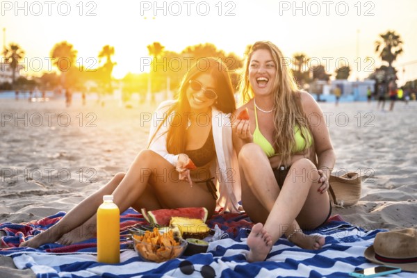 Two female friends enjoying a picnic with fresh fruit and juice on a beach towel at sunset,