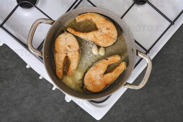Salmon fillets are being cooked in a pan with garlic on a stovetop. The kitchen is well-lit and the
