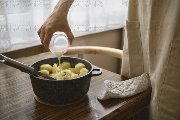 A hand pours warm milk onto a pot filled with boiled potatoes on a wooden table. The process of