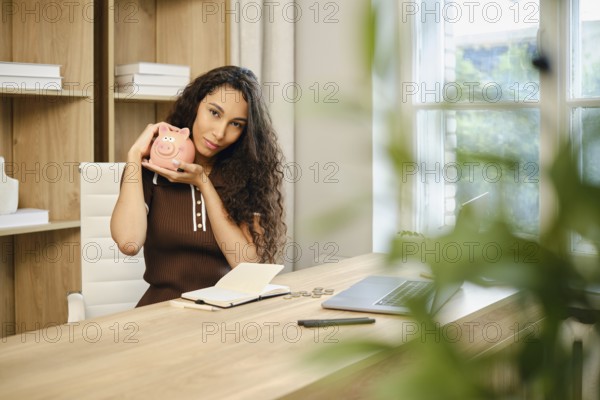 In a contemporary office space, a woman with curly hair sits at a wooden desk, smiling as she holds