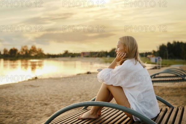 A woman sits comfortably on a wooden bench beside a serene lake, gazing at the vibrant sunset. The